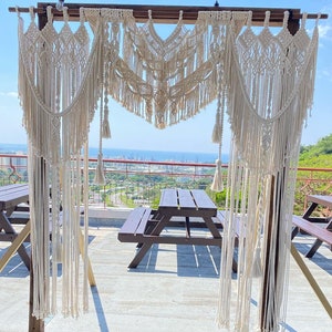 Op de afbeelding: Een witte macramé boog met kwastjes hangt aan een houten frame. De boog is buiten op een terras opgezet met twee houten picknicktafels op de voorgrond.