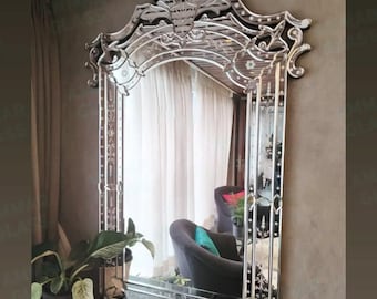 May include: A large, ornate mirror with a decorative, etched frame. The mirror has a rectangular shape with an arched top and intricate floral and geometric designs. The frame is silver and the mirror reflects a room with a plant and a dark gray armchair.