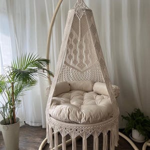 Op de afbeelding: Een beige macramé hangstoel met een ronde, gewatteerde zitting en rugleuning. De stoel heeft een gefranjerde onderkant en is opgehangen aan een gebogen houten frame. Decoratieve kussens en een potplant zijn zichtbaar.