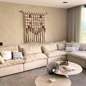 May include: A beige sectional sofa with multiple throw pillows in a living room setting. A decorative wall hanging with white and colorful accents hangs above the sofa. A round coffee table with decorative objects and a black fan is in front of the sofa.