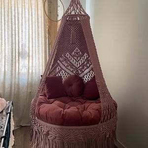 May include: A hanging macrame chair in a dusty rose color. The chair features a round, plush cushion and three matching pillows. The chair is suspended from the ceiling and has a fringed bottom.