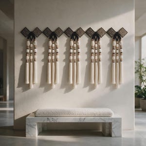 May include: A wall-mounted decorative piece featuring five black diamond-shaped panels with geometric patterns, each suspending three long cream-colored fiber tassels accented with wooden beads and black cord loops, positioned above a white marble bench.