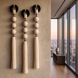 May include: Three vertical wall hangings featuring a series of stacked off-white fabric spheres separated by dark grey cylindrical spacers. Each piece is topped with a black circular mount and terminates in a long, flowing off-white tassel.