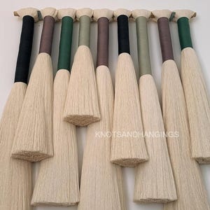 May include: A set of ten white fluffy tassels with different colored trim, hanging from a wooden dowel. The tassels are all the same size and shape, and they are arranged in a row. The trim colors are black, brown, green, and blue.