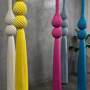 May include: A collection of colorful, hanging decorative tassels. The tassels are in shades of white, yellow, pink, blue, and gray. Each tassel features a knotted sphere design and a long, fringed bottom, creating a modern, artistic display.