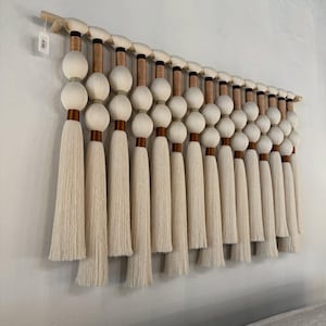 May include: A wall hanging featuring a horizontal wooden rod with multiple suspended off-white yarn elements. Each element consists of two stacked spheres wrapped in off-white and copper-colored thread, ending in a long, straight fringe tassel.