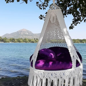 Puede incluir: Una silla colgante de macramé blanco con flecos en la parte inferior y un cojín de terciopelo morado. La silla está suspendida de una rama de árbol con vistas a un lago y una cordillera. El diseño de la silla presenta intrincados nudos.