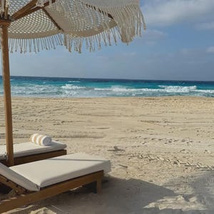 Puede incluir: Una escena de playa con una sombrilla blanca con flecos, tumbonas de madera y una toalla enrollada. El océano turquesa se encuentra con la playa de arena bajo un cielo azul con nubes. La escena evoca relajación y ocio.