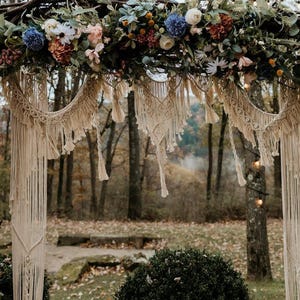 Peut inclure: Une arche de mariage décorée d'une guirlande florale de différentes couleurs, notamment des fleurs bleues, roses et blanches. L'arche est ornée de suspensions en macramé, créant une ambiance bohème et rustique.