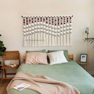 May include: A white macrame wall hanging with red and white stripes and white pom poms. The wall hanging is above a green bed with white pillows and a white blanket. The text "KNOTSANDHANGINGS" is visible on the wall hanging.
