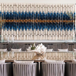 May include: A large, woven wall hanging with a pattern of white, blue, and gold. The wall hanging is above a dark wood cabinet. Below, a dining table is set with chairs with gray ribbon-like backs.
