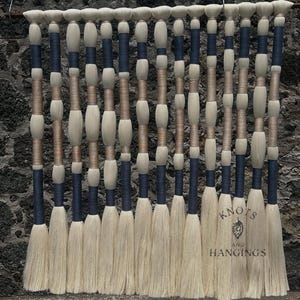 May include: A decorative wall hanging featuring vertical strands of natural-colored fibers. The design incorporates navy blue and tan accents, with a textured appearance. The bottom of the strands have a fringed effect. The text "Knots and Hangings" is visible.