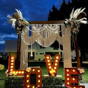 Puede incluir: Un arco de boda de madera con decoración colgante de macramé color crema y arreglos florales, situado detrás de grandes letras luminosas que forman la palabra LOVE, decoradas con follaje verde y pequeños ramos de flores.