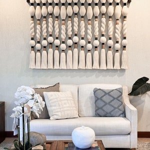 May include: A white macrame wall hanging with a wooden frame and tassels. The wall hanging is made of white and beige yarn and has a geometric pattern. The wall hanging is hanging on a white wall above a white couch with pillows.
