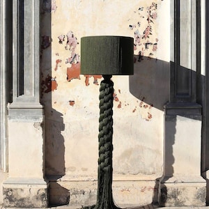 May include: A floor lamp with a dark green lampshade and a braided rope-like base. The base has a tassel at the bottom. The lamp is set against a textured wall with a distressed appearance.