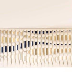 May include: A cream-colored wall hanging with a wooden rod and black brackets. The design features white pom-poms, gold and navy blue vertical accents, and white tassels at the bottom. This decorative piece adds texture and visual interest to any space.