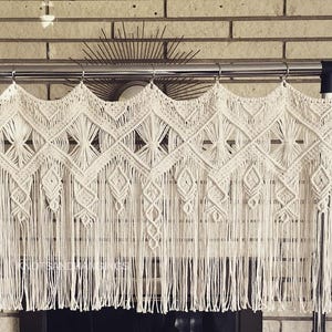 May include: A white macrame curtain with a geometric pattern hangs from a metal rod. The curtain is made of thick, knotted cords and has a fringe at the bottom.
