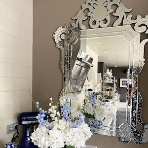 Pode incluir: Espelho biselado ornamentado com desenhos florais e de rolagem gravados. O espelho reflete uma cena de cozinha com arranjos florais brancos e azuis e uma batedeira azul. O texto "KNOTS&HANGINGS" é visível.