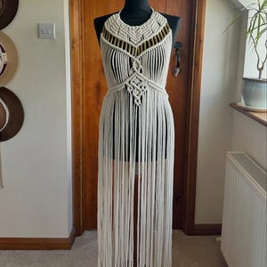May include: A cream macrame dress on a mannequin. The dress has a halter neckline with gold detailing and a detailed bodice. Long fringe hangs from the waist to the floor. The dress is a bohemian style, suitable for a summer occasion.