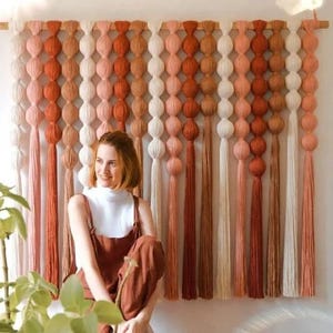 May include: A large, handmade wall hanging featuring a gradient of colors from white to rust. The design includes rows of large, spherical knots and long tassels. The piece is mounted on a wooden dowel, creating a bohemian, textured wall decor.