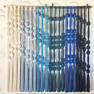 May include: A macrame wall hanging featuring a gradient of colors from cream to navy blue. The design includes knotted sections and tassels, creating a textured, decorative piece. The wall hanging is mounted on a wooden dowel.