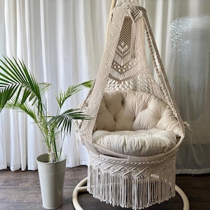 May include: A cream-coloured macrame hanging chair with a buttoned cushion. The chair has a fringe at the base and is suspended from a wooden bar. A potted plant is to the left.