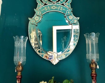 May include: A large, ornate, oval-shaped mirror with a silver frame and etched floral designs. The mirror is hanging on a teal wall with two decorative candle holders on either side. The candle holders are made of glass and have a red and white floral design.