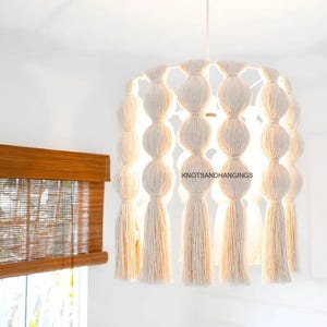 May include: A white macrame light fixture with a round shape and tassels hanging from the bottom. The light fixture is lit and the light shines through the macrame. The text "KNOTSANDHANGINGS" is visible on the fixture.