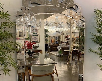 May include: A large, ornate mirror with an arched top and etched floral and scroll designs. The mirror reflects a room filled with chandeliers and furniture. The text "KNOTS&HANGINGS" is visible on a chair.