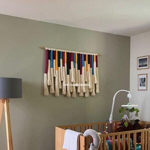 May include: A white, cream, yellow, blue, and burgundy macrame wall hanging with the text "KnotsAndHangings" in the center. The wall hanging is in a nursery with a crib and a mobile.
