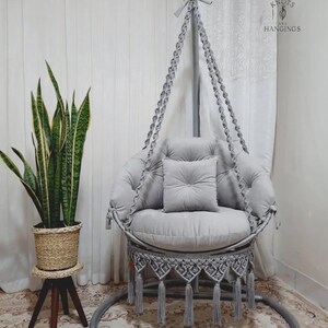 May include: A grey macrame hanging chair with a round seat and backrest, featuring a decorative fringe and tassels. The chair is suspended from a metal frame and includes a matching cushion and pillow. A potted plant is to the left.