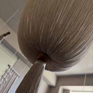 Pode incluir: Uma grande luminária pendente de macramê bege. A luminária tem uma forma arredondada com uma longa franja em forma de borla na parte inferior. A luminária é suspensa do teto, mostrando uma estética natural e boêmia.