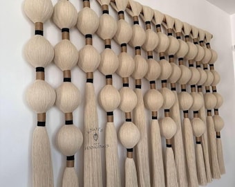 May include: A large, cream-colored wall hanging with a wooden rod. The design features multiple rows of large, spherical shapes with black and brown accents, and long, dangling tassels. The piece is mounted on a white wall.