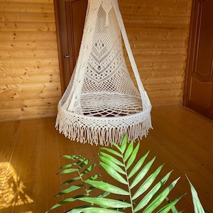 May include: A cream macrame hanging chair with a fringed base and a pointed top. The chair is suspended from the ceiling in a room with wooden walls and flooring. Green foliage is in the foreground.