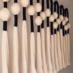 May include: A wall hanging featuring a series of decorative elements. Each element has a black, curved top, a white sphere, a black cylindrical section, and a long, white, fringed bottom. The arrangement creates a visually appealing, textured display.