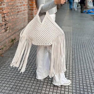 Może przedstawiać: Biała torba typu tote wykonana na szydełku z długimi frędzlami. Torba wykonana jest z grubego sznurka bawełnianego i ma szeroki, otwarty górny brzeg.