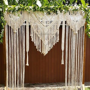 May include: A white macrame wedding arch with a wooden frame. The arch is decorated with a variety of macrame designs, including a large diamond-shaped piece in the centre. The arch is set up outdoors against a brown wooden fence.