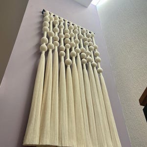 May include: Cream-colored macrame wall hanging with a series of large, round beads and long, flowing tassels. The wall decor is mounted on a light purple wall, creating a neutral and textured aesthetic. The piece adds a bohemian touch to any space.