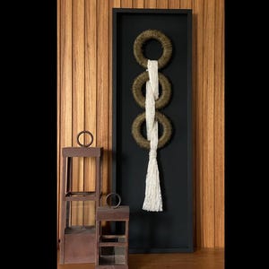 May include: A vertical black framed wall hanging with three woven rings in a chain, connected by white rope. Two small, brown, wooden side tables with a circular metal ring on top are in the foreground. The background is a wooden wall.