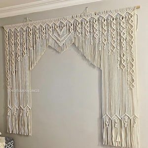May include: A white macrame wall hanging with a geometric design. The hanging is made of cotton cord and has a fringe at the bottom. The hanging is attached to a wooden dowel rod.