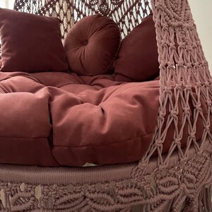 May include: A hanging macrame chair in a muted rose color. The chair features a plush, tufted seat and several matching pillows. The macrame design is visible on the sides and top, with a fringe detail at the bottom. The overall aesthetic is bohemian and cozy.