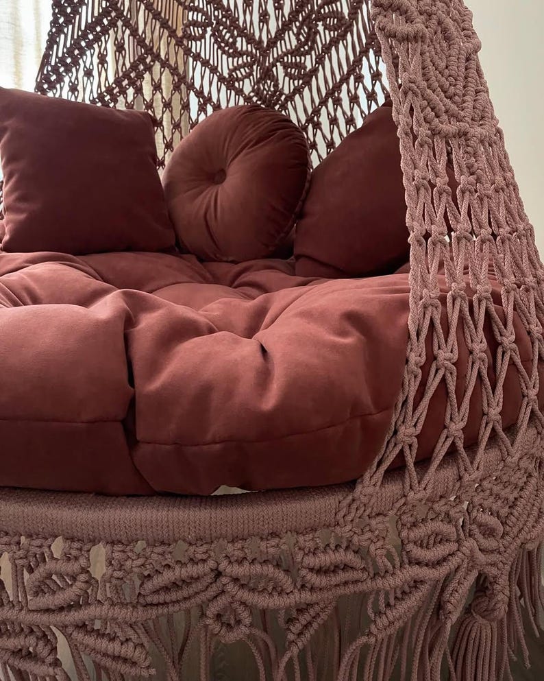 May include: A hanging macrame chair in a muted rose color. The chair features a plush, tufted seat and several matching pillows. The macrame design is visible on the sides and top, with a fringe detail at the bottom. The overall aesthetic is bohemian and cozy.