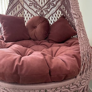 May include: A hanging macrame chair in a dusty rose color. The chair features a large, round, tufted cushion and three matching pillows. The macrame design is visible on the back and sides of the chair, with a fringe detail at the bottom.