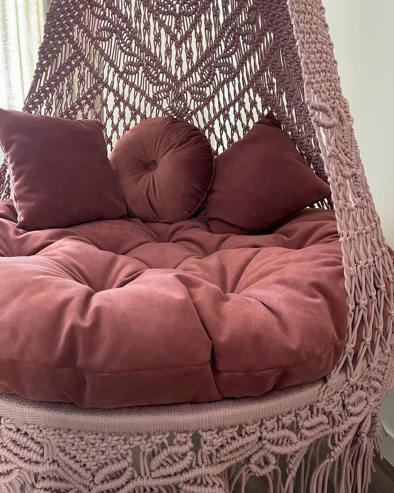 May include: A hanging macrame chair in a dusty rose color. The chair features a large, round, tufted cushion and three matching pillows. The macrame design is visible on the back and sides of the chair, with a fringe detail at the bottom.