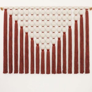 May include: A wall hanging featuring a design of white spheres and rust-colored tassels. The spheres are arranged in rows, and the tassels hang vertically. The piece is mounted on a natural wood rod, creating a modern, textured wall decor.