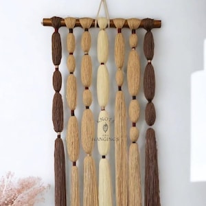May include: A handcrafted wall hanging featuring a wooden rod with various lengths of yarn in shades of brown, beige, and cream. The yarn is knotted and arranged in a vertical pattern, with the text "Knots and Hangings" visible.
