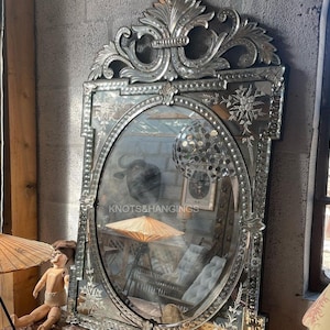 May include: A large, ornate silver mirror with a beveled oval frame. The frame is decorated with intricate floral designs and a beaded border. The mirror reflects a room with furniture and a decorative umbrella.