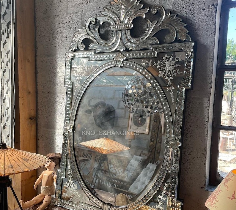May include: A large, ornate silver mirror with a beveled oval frame. The frame is decorated with intricate floral designs and a beaded border. The mirror reflects a room with furniture and a decorative umbrella.