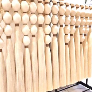 Pode incluir: Uma coleção de tapeçarias de parede em macramé branco sujo. Cada peça apresenta uma série de grandes contas de madeira arredondadas com detalhes castanhos, que conduzem a uma longa borla fluida. As tapeçarias são exibidas numa prateleira de metal preto.
