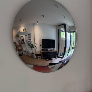 May include: A large, circular, frameless wall mirror with a reflective surface that creates a wide-angle, convex-style distortion of the room it faces, showing a living area with a television, furniture, and indoor plants.